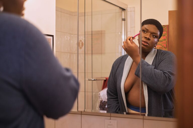 A Non Binary Trans Woman Applying Makeup In A Mirror.jpg