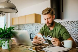 Man Sitting In Front Of Laptop Holding Credit Card While Using Smart Phone While Working From Home Izusek.jpg